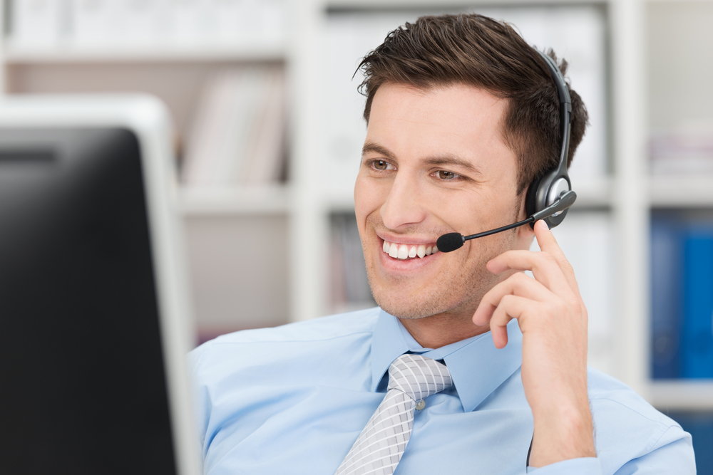 Smiling friendly handsome young male call centre operator or client services personnel beaming as he listens to a call and checks information on his computer monitor Smiling friendly handsome young male call centre operator or client services personnel beaming as he listens to a call and checks information on his computer monitor